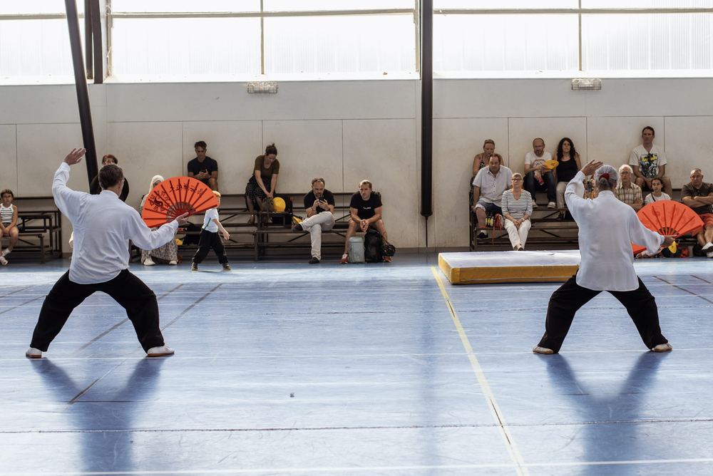 Deux pratiquants de Tai Chi Chuan face à face, éventail rouge à la main, devant le public du gala